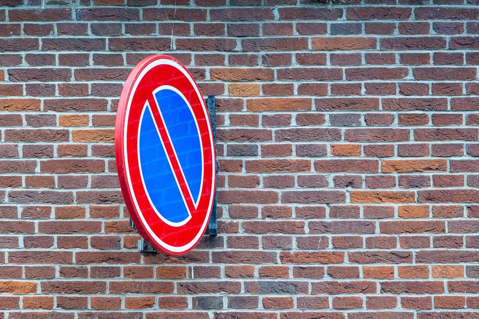 A circular no parking sign mounted on a metal bracket, attached to a red brick wall. The sign has a blue background with a red border and a red diagonal line crossing through the centre, indicating no parking or stopping. The brick wall behind the sign features a traditional running bond pattern with reddish-brown bricks and contrasting mortar joints. The setting appears to be outdoors, likely on a street or building frontage, with the sign positioned at eye level. This visual detail is relevant to house removals and transport, highlighting parking restrictions that may impact the home relocation process, as managed by Man with Van Beckton. The overall scene emphasizes the importance of understanding local parking rules during furniture transport and loading activities, especially in coordination with removal services in the Newham area.