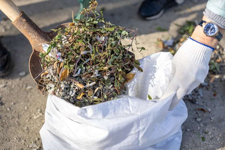A person wearing a white glove and a wristwatch is using a shovel to transfer a mixture of dried leaves, small twigs, and gravel into a large white removal bag. The bag is positioned on a gravel or di