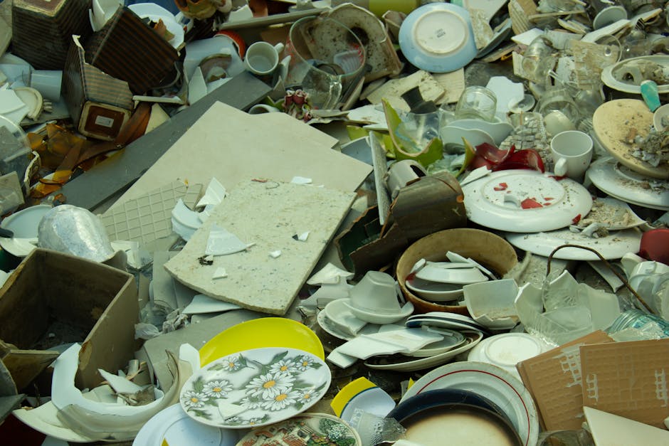 The image displays a large pile of discarded household items and waste materials, including broken and intact ceramic dishes, cups, plates, and bowls, some with visible cracks or chips. There are also cardboard boxes, plastic containers, and packaging materials scattered across the pile, with a few pieces of fabric or cushioning material among the debris. The items are placed on a paved surface outside a property, suggesting a waste disposal or clearance scene related to home relocation or removal services. In the background, a portion of a property entrance or driveway is visible, supporting the context of house removals, as offered by Man with Van Beckton, with the focus on the disposal of bulky waste during a moving process.