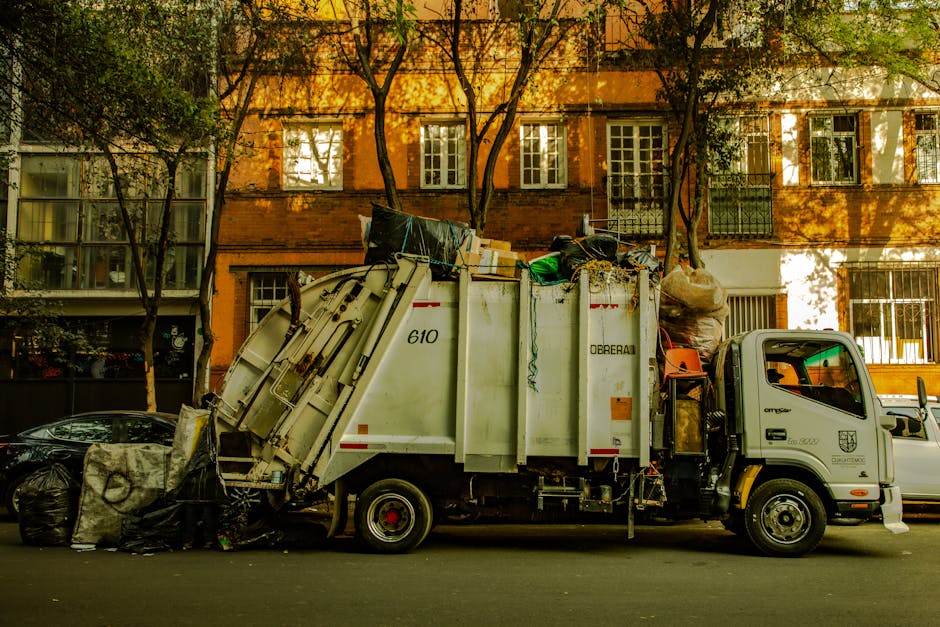 A white garbage collection truck is parked on a street in Beckton, loaded with various waste materials including black plastic bins, cardboard boxes, and large bags of rubbish. The truck is positioned beside a row of multi-storey residential buildings with large windows, some of which have white frames and security bars. The truck's cargo area is filled with mixed bulky waste, and the rear is tilted downward as if in the process of collecting or dumping waste. Nearby, a black car is also parked, partially obscured by the waste bins. The scene is illuminated by natural daylight, with shadows cast by trees and buildings, and the pavement area around the truck is clear for loading. This image, associated with home relocation and waste disposal services by Man with Van Beckton, illustrates the process of waste removal during a house move or property clearance, involving equipment like waste bins and collection vans.