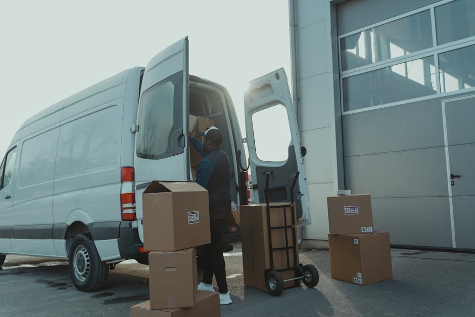 A professional mover from Man with Van Beckton is engaged in a home relocation, standing outside a modern building with large glass windows, unloading various packed cardboard boxes from a light grey 