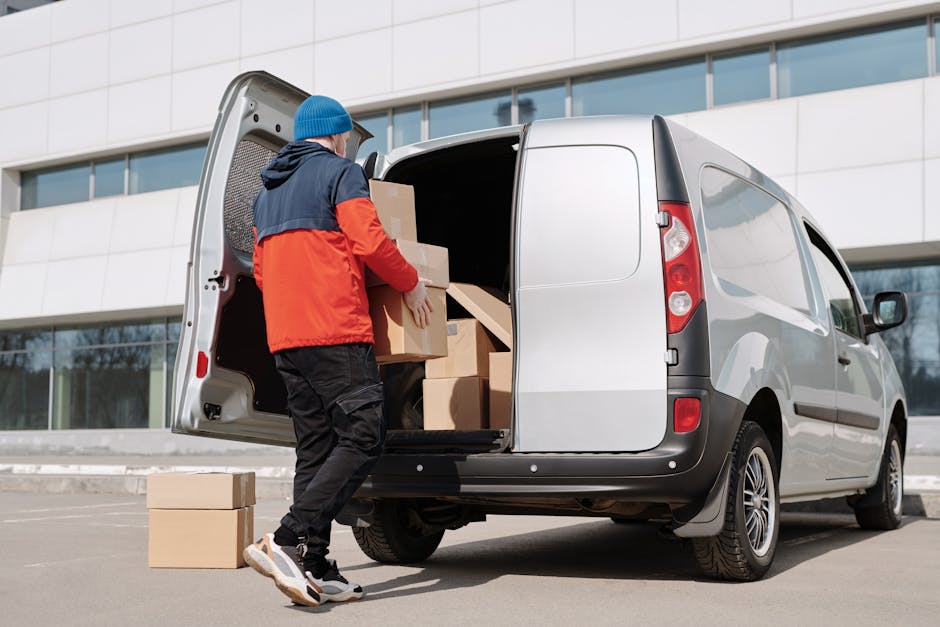A professional mover from Man with Van Beckton is engaged in a home relocation, standing outside a modern building with large glass windows, unloading various packed cardboard boxes from a light grey 