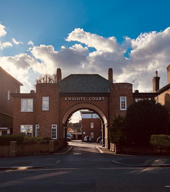 The image depicts the entrance to Knights Court, a residential building constructed from red brick, featuring an archway with the name 'KNIGHTS COURT' inscribed above. On either side of the arch, there are two small towers with rounded tops, also made of red brick. To the left and right of the entrance, there are multiple white-framed windows, some of which are visible on the two-story building. The driveway leading through the archway appears to be part of a property managed by Man with Van Beckton, as indicated by the context, and is bordered by low brick walls and well-maintained bushes. Behind the arch, a parking area with several vehicles is visible, along with additional residential buildings in the background. The sky above is partly cloudy with patches of sunlight illuminating parts of the scene, suggesting a clear day, suitable for house removals or moving activities. This setting reflects an urban residential environment associated with property relocation and furniture transport services.