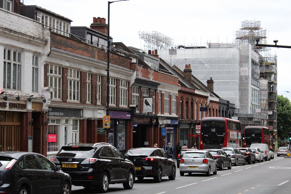 A city street scene during daytime showing a row of brick and stucco terraced houses with large windows; some buildings are undergoing renovation, covered in scaffolding and white protective sheeting. In the foreground, there is a line of parked cars including sedans and hatchbacks, with a few pedestrians walking along the pavement. Two red double-decker buses are visible, one approaching and one parked at a bus stop. The street is busy with vehicles in motion and parked along the curb, and the scene is lit with natural daylight. The image captures an urban environment typical of a home relocation or moving service context, with potential for furniture transport and loading activities linked to house removals, as operated by Man with Van Beckton.