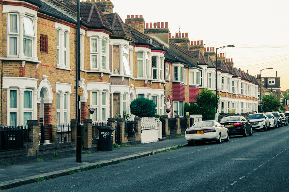 A row of traditional terraced houses with brick exteriors and bay windows, situated along a residential street in Beckton. Front gardens are enclosed by low brick walls and black metal fencing, with some featuring small trees, shrubs, and decorative elements. The pavement in front of the houses has an array of parked cars, including sedans and hatchbacks, lined up along the curb. A black wheelie bin marked 'DOGG' stands near one of the fences, close to a black utility pole with a yellow box attached. Streetlights and overhead electrical wires are visible, with gentle daylight illuminating the scene under a clear sky. This neighborhood showcases characteristic Victorian-style architecture with multiple chimneys on rooftops, reflecting typical house designs in Beckton, suitable for home relocation or furniture transport services such as those offered by Man with Van Beckton during house removals or moving processes.