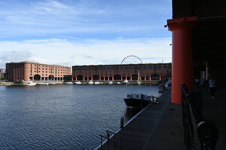 A view of a waterfront area near Royal Albert Dock in Beckton, showing a large red brick warehouse-style building with multiple arches along the water's edge. Several small boats are moored close to the dock, with one dark-colored vessel tied to the wooden pier in the foreground. The image captures an outdoor scene with a partly cloudy sky above, and a Ferris wheel faintly visible in the distance behind the building. On the right side, part of a covered walkway with red support columns and black metal railings is visible, with a few people walking along the path. The scene reflects typical elements involved in home relocation and furniture transport, such as packing materials and the loading process of moving items onto boats or vehicles while adjacent to a water-based transportation hub. Occasionally, Man with Van Beckton provides removals and moving services in this area, facilitating efficient packing and house removals near iconic docks and waterways.