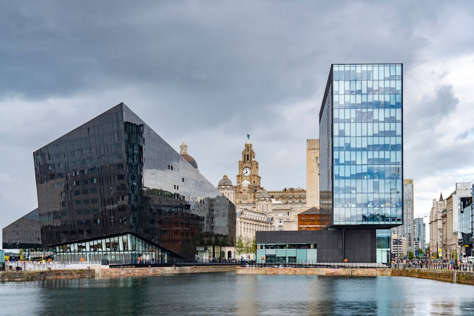 The image depicts a modern urban scene showing the waterfront with two distinctive contemporary office buildings, one with a reflective black glass facade and the other with a blue-tinted glass exterior, positioned on the edge of a canal or river. In the background, there are historic buildings, including a clock tower with a green dome and ornate architecture, set against a cloudy sky. The foreground features a paved area near the water, possibly part of a pedestrian walkway or loading zone. Although no specific moving activity is visible, the setting reflects the type of cityscape where house removals and furniture transport services like those provided by Man with Van Beckton might operate, especially when coordinating home relocation logistics near central Liverpool.