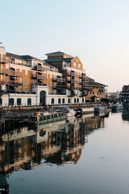 A view of a waterfront area near Royal Albert Dock in Beckton, showing a large red brick warehouse-style building with multiple arches along the water's edge. Several small boats are moored close to the dock, with one dark-colored vessel tied to the wooden pier in the foreground. The image captures an outdoor scene with a partly cloudy sky above, and a Ferris wheel faintly visible in the distance behind the building. On the right side, part of a covered walkway with red support columns and black metal railings is visible, with a few people walking along the path. The scene reflects typical elements involved in home relocation and furniture transport, such as packing materials and the loading process of moving items onto boats or vehicles while adjacent to a water-based transportation hub. Occasionally, Man with Van Beckton provides removals and moving services in this area, facilitating efficient packing and house removals near iconic docks and waterways.