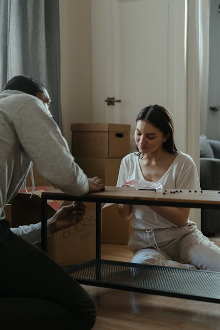Two individuals are indoors with moving boxes visible behind them, suggesting a home environment during a relocation process. One person, seated on the floor wearing white casual clothing, is looking down at a small object in their hands. The other person, standing and wearing a grey hoodie, is leaning over the edge of a moving trolley or dolly, engaging with the seated individual. The trolley appears to be used for transporting smaller items or packing materials. In the background, there are cardboard boxes, some labeled with handwritten text, stacked near a door and a window with curtains, indicating preparations for a move in a property with natural light coming through. The scene captures the packing and moving phase typical of house removals or furniture transport, with visible packing materials like cardboard boxes and the use of practical equipment such as the trolley. Man with Van Beckton provides such services, ensuring careful handling of furniture and belongings during home relocations in the Beckton area.
