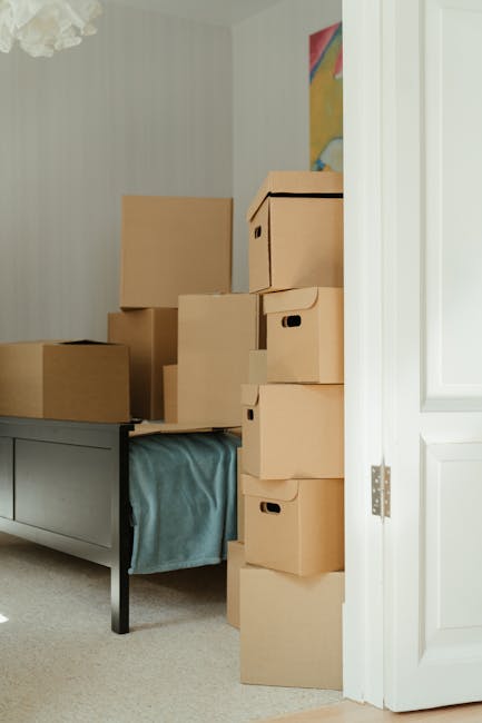 Inside a room with light-colored walls and a gray-painted wooden bed frame, several stacked cardboard boxes of various sizes are positioned along the wall and partially on the bed, indicating a packing or moving process. Some boxes are sealed with packing tape, while others are open or partially closed, revealing no contents. The boxes are arranged in a vertical stack, with larger ones at the bottom and smaller ones on top, suggesting preparation for a home relocation. Natural light illuminates the space, highlighting the clean environment. To the right, a partially open white door with decorative paneling provides access to the room. This scene illustrates the packing and loading activities associated with professional removals services, such as those offered by Man with Van Beckton, during furniture transport or home moving procedures.