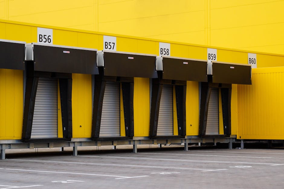A row of four loading bays at a commercial warehouse or storage facility, painted in bright yellow with each bay numbered from B56 to B60, equipped with black retractable dock shelters and grey rollin