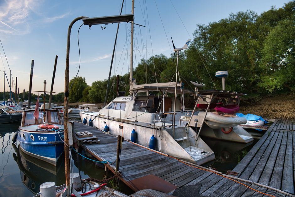 A marina with numerous small boats and narrowboats moored along wooden docks on calm water, surrounded by modern multi-storey residential buildings with balconies and large windows, under a bright blu