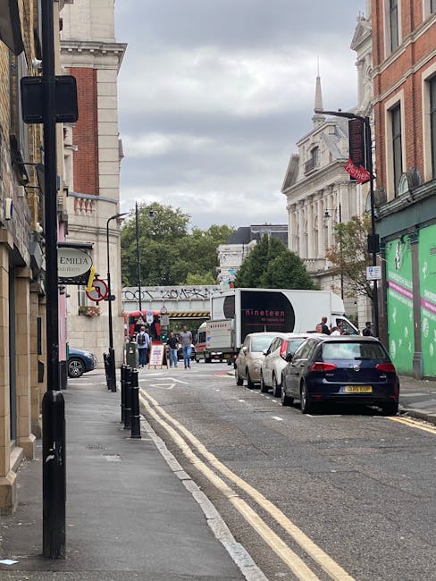 A narrow street in Beckton with parked cars lining both sides, including a black car closest to the camera. On the right side, a green storefront is visible, while the left side features a sign for 'Emilia.' In the distance, a white moving truck with the black and red 'Dino' logo is being loaded or unloaded, with several people around the vehicle. The street has black bollards on the pavement, and overhead, there is a pedestrian bridge with some graffiti on it. The sky is overcast, and the surrounding buildings are a mix of classical and modern architecture, including a prominent white building with columns and a clock tower at the end of the street. This scene depicts urban logistics typical of a house or flat relocation process, possibly involving furniture transport and moving activities, coordinated by a professional company such as Man with Van Beckton, in a setting suitable for home removals and packing and moving services.