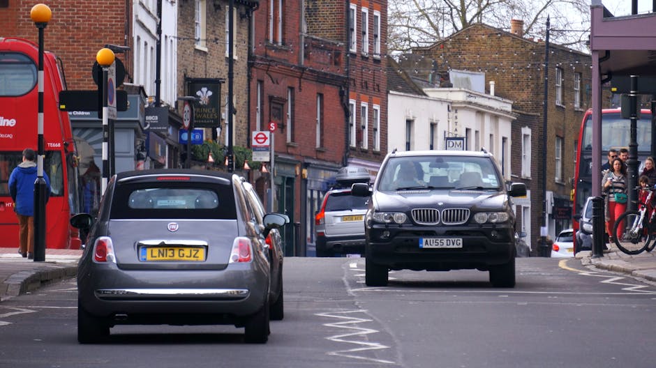 A street scene on South Beckton Street showing parked vehicles including a small silver Fiat 500 with a yellow UK registration plate and a black BMW SUV with a white UK registration plate, both positioned on the curbside. Behind these, additional cars are parked along the narrow, urban street with brick and white-painted buildings lining the sides. Pedestrians are visible walking on the pavement, some near bus stops with covered shelters. The street is shaded and appears busy, with a red double-decker bus partially visible on the left side, indicating active street traffic. The environment suggests a typical residential and commercial area suitable for home relocations. Man with Van Beckton specializes in house removals and furniture transport, and this scene illustrates the challenges of loading and navigating narrow streets during a house move, especially on busy days.