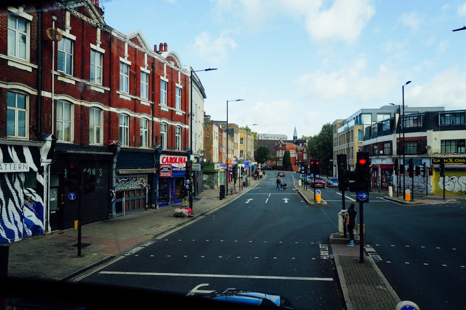 A narrow street in Beckton with parked cars lining both sides, including a black car closest to the camera. On the right side, a green storefront is visible, while the left side features a sign for 'Emilia.' In the distance, a white moving truck with the black and red 'Dino' logo is being loaded or unloaded, with several people around the vehicle. The street has black bollards on the pavement, and overhead, there is a pedestrian bridge with some graffiti on it. The sky is overcast, and the surrounding buildings are a mix of classical and modern architecture, including a prominent white building with columns and a clock tower at the end of the street. This scene depicts urban logistics typical of a house or flat relocation process, possibly involving furniture transport and moving activities, coordinated by a professional company such as Man with Van Beckton, in a setting suitable for home removals and packing and moving services.