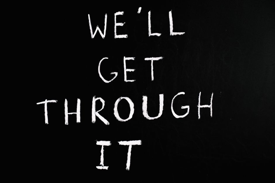 A black chalkboard with the phrase 'We'll Get Through It' written in large, white chalk letters arranged vertically. The background is plain black, and the message is centered on the board, suggesting resilience and encouragement. This image does not depict furniture, boxes, or moving equipment. It may be used in the context of home relocation or moving services provided by Man with Van Beckton, emphasizing support during the packing and moving process associated with house removals or furniture transport, especially relevant to understanding or managing unexpected charges in quotes for removals.
