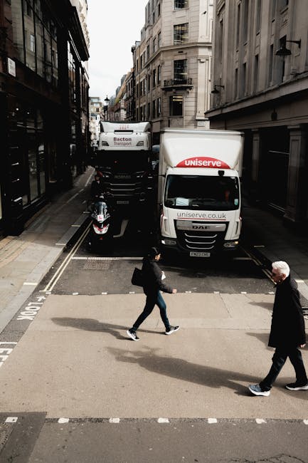 A street scene showing two vehicles involved in house removals, with one being a white moving van marked with 'uniserve' and a smaller white vehicle parked behind it. The van is positioned near the curb on a narrow, urban street lined with multi-storey buildings on both sides. In front of the vehicles, two pedestrians are crossing the street, one carrying a black bag and the other with white hair and glasses, both dressed in dark clothing. The pavement has striped markings indicating pedestrian crossing areas, and the scene is lit by natural daylight with overcast skies visible. This setting illustrates the logistics of furniture transport and home relocation services provided by [COMPANY_NAME], capturing the loading process or street-based moving activity typical of urgent same-day removals in Beckton.