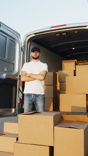 A man wearing a dark cap, white t-shirt, and jeans stands with his arms crossed in front of an open van, positioned on a paved surface outdoors. The back of the van is filled with neatly stacked cardboard boxes of various sizes, some of which are partially unwrapped or open, showing packing materials like plastic wrap and paper. Additional boxes are placed on the ground nearby, with a few open, revealing their contents. The interior of the van is illuminated, highlighting the boxes and equipment such as trolleys and straps used during house removals. The scene depicts a relocation or furniture transport process, likely part of a professional house moving service, with a focus on loading or unloading items for home relocation, as part of a same-day removals service in Beckton. Man with Van Beckton is a reliable provider handling the logistics of packing and moving within this environment.